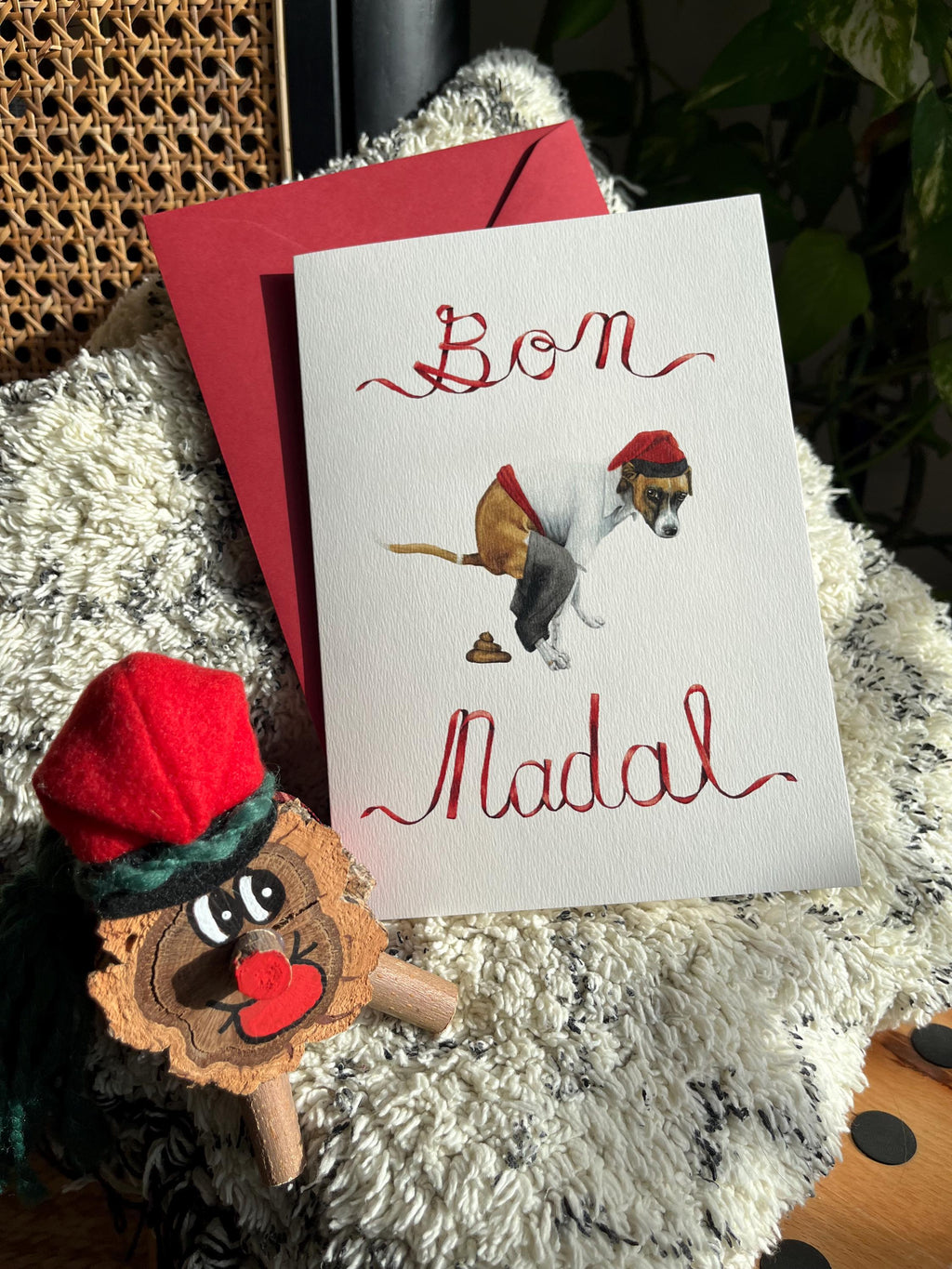 custom caganer christmas Card handpainted in watercolor with 'Bon Nadal' ribbon text and a painting of a dog pooping while wearing the traditional catalan farmers clothes, accompanied by a small wooden tio de nadal figurine on a textured surface.
