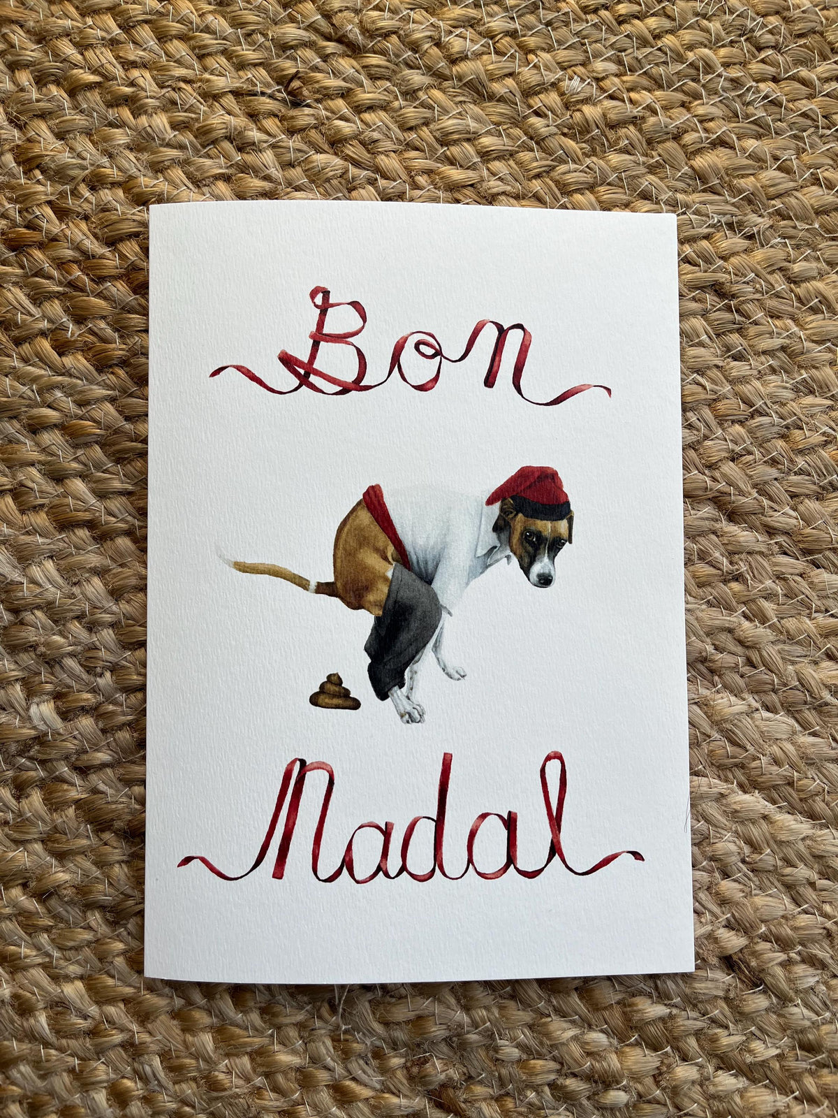 handpainted watercolor illustration of a ratonero bodeguero dog pooping wearing a traditional catalan farmers outfit in the style of a caganer. above and below it is the text "Bon Nadal" written in ribbon