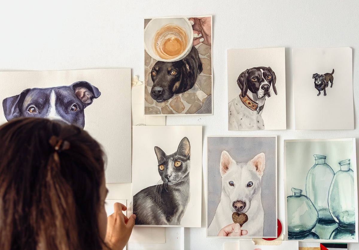 the back of a woman's head as she adjusts a painting on a white wall full of handpainted watercolor pet portraits. There are 5 dog paintings, 1 cat painting and a painting of three glass bottles.